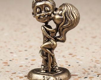 Puede incluir: Figurita de bronce de una pareja abrazándose y besándose. La figura masculina sonríe, mientras que la femenina le da un beso en la mejilla. La base en forma de corazón tiene grabados los números "13" y "14".