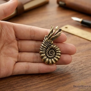 Könnte beinhalten: Eine detaillierte Ammoniten-Skulptur aus Messing mit einer Spiralmuschel und tentakelartigen Merkmalen. Das Fossil im antiken Stil ist ein dekoratives Objekt, das aufwendige Handwerkskunst und eine Vintage-Ästhetik zeigt. Holz Hintergrund.