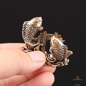 May include: A bronze-colored decorative figurine featuring two fish swimming towards each other, separated by a small archway. The fish have detailed scales and are set on a base with wave-like patterns. The figurine is held in a hand.
