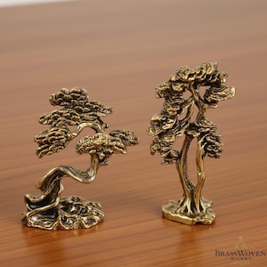 May include: Two small, detailed brass bonsai tree sculptures. One tree has a curved trunk and the other is upright. The trees are on a wooden surface. The logo "BrassWoven Works" is in the bottom right corner.