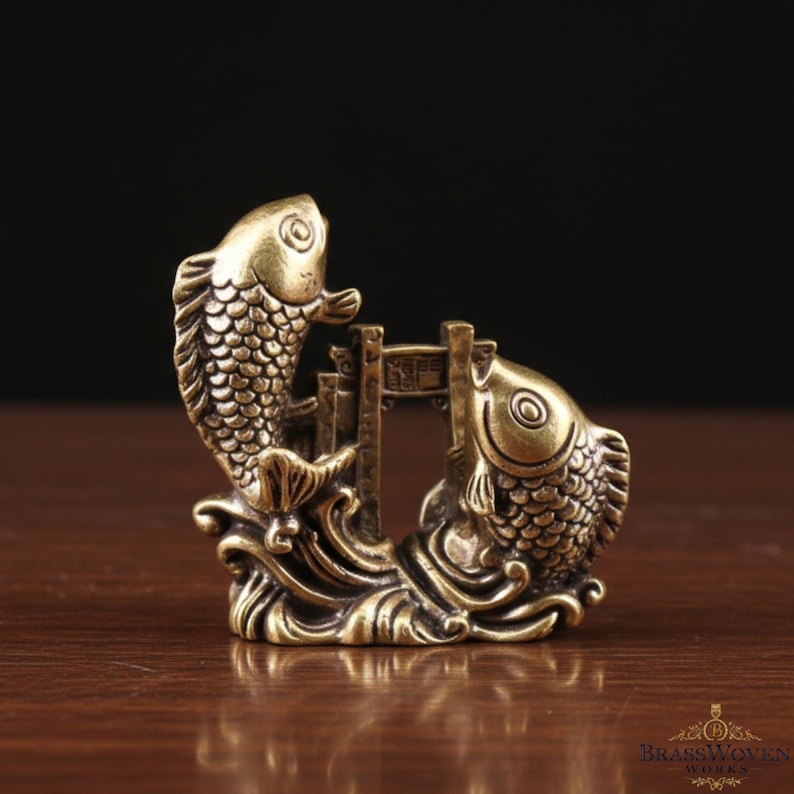 May include: A brass sculpture featuring two stylized fish swimming towards each other, with a small bridge between them. The fish have detailed scales and fins, and the sculpture sits on a wave-like base. The overall design is intricate and decorative.