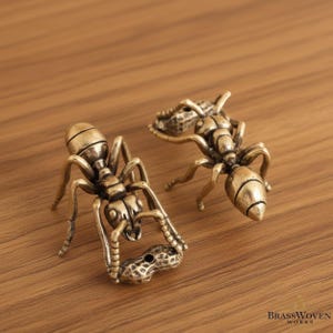 May include: Two detailed brass ant figurines, each holding a peanut. The ants are a warm gold color, with intricate details on their bodies and legs. The peanuts are also brass, with a textured surface. The figurines are on a wooden surface.
