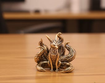 May include: A detailed brass fox figurine with multiple tails, sitting upright. The fox has pointed ears and intricate detailing on its body. The figurine is on a wooden surface.