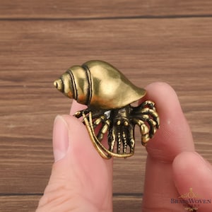 May include: A detailed brass figurine of a hermit crab. The crab has a textured shell and intricate detailing on its body and claws. The figurine is held in a person's hand, showcasing its size and craftsmanship. The overall design is a realistic representation.