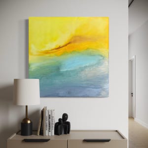 May include: Abstract art piece with a vibrant color palette. The artwork features a gradient of yellow, orange, blue, and navy hues, evoking a sense of a sunset or seascape. The painting is displayed above a cabinet with a lamp, books, and a sculpture.
