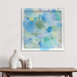 May include: Abstract art print in a white frame featuring blue, green, and yellow watercolor-style brushstrokes. The artwork is displayed above a wooden table with two white ceramic vases.