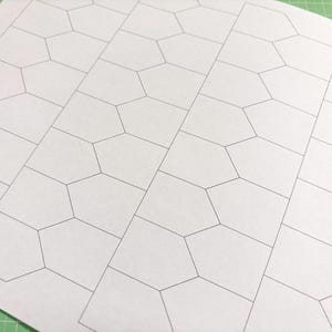 Könnte beinhalten: Ein weißes Blatt Papier mit einem sich wiederholenden geometrischen Muster aus verbundenen Fünfecken, umrandet von dünnen schwarzen Linien. Das Muster ist in diagonalen Reihen angeordnet und erzeugt einen texturierten Effekt. Das Papier liegt auf einer grünen Schneidematte.