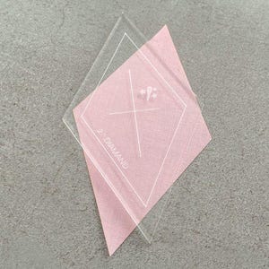 Könnte beinhalten: Eine transparente, rautenförmige Quiltvorlage liegt auf einem rosafarbenen Stoffdiamanten. Die Vorlage hat weiße Markierungen und den Text "2" und "DIAMOND". Der Stoff hat ein Kreuzstichmuster.