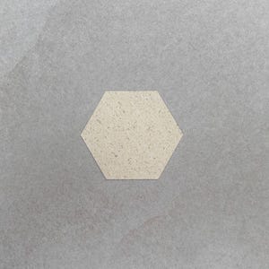 Könnte beinhalten: Ein sechseckiger Untersetzer mit einer gesprenkelten, cremefarbenen Oberfläche. Der Untersetzer besteht aus einem strukturierten Material und ist vor einem grauen Hintergrund platziert. Dieser Artikel eignet sich für die Heimdekoration.