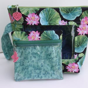 May include: A large zippered project bag with a black background featuring pink lotus flowers and green lily pads, paired with a smaller square zippered pouch in a textured teal pattern. Both bags have clear vinyl windows and pink heart-shaped zipper pulls.