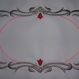 May include: A decorative embroidery design with a pink oval frame and two stylized floral motifs in the top and bottom corners. The floral motifs are in shades of brown, green, and red.