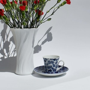 May include: A white ceramic vase with a vertical wavy texture holds a bouquet of red carnations. Beside it sits a white porcelain teacup and saucer featuring a repeating blue floral pattern on the exterior and the matching saucer surface.