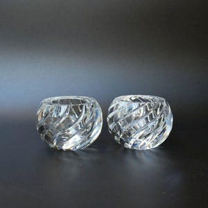 May include: Two clear glass votive candle holders. Each has a round shape with a spiral design carved into the glass. The candle holders are on a dark background.