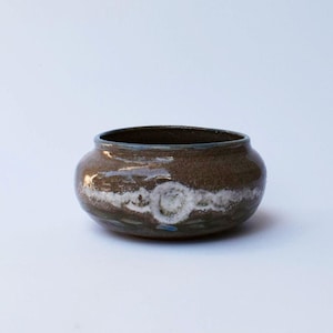 May include: A small, round, brown ceramic bowl with a glossy finish. The exterior features a horizontal band of textured, mottled white glaze that encircles the middle, creating a distinct contrast against the dark, earthy brown clay body.