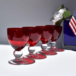 May include: A set of four matching wine glasses featuring deep red glass bowls and clear glass stems with a spherical accent above the circular base. The glasses are arranged in a diagonal line, casting distinct red shadows on the surface.