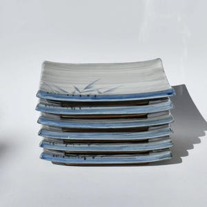 May include: A stack of rectangular ceramic plates. Each plate has a light gray background with blue accents along the edges. A subtle bamboo design is painted on the top of each plate.