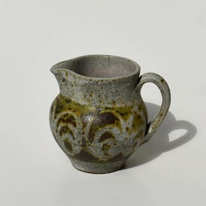 May include: A small, handcrafted ceramic creamer with a rounded body and a handle. The creamer features a speckled gray and green glaze with brown decorative patterns. The spout is small and the interior is a light gray color.