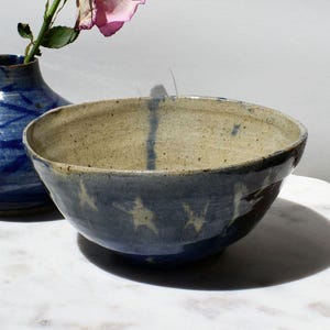 May include: A handmade ceramic bowl with a speckled beige interior and a dark blue exterior. The bowl features white star-like designs and a dark blue vertical stripe. A blue vase and a pink flower are in the background.