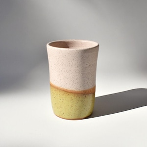 May include: A cylindrical ceramic tumbler featuring a speckled finish. The upper portion is a pale, creamy off-white, while the lower section is a vibrant chartreuse yellow. The two colors meet in a wavy, horizontal line around the vessel.