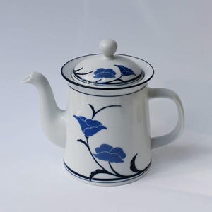 May include: White ceramic teapot with a lid, featuring a blue floral design. The teapot has a curved spout and a handle. The lid is adorned with a small knob. Black trim accents the rim and base, creating a classic aesthetic.