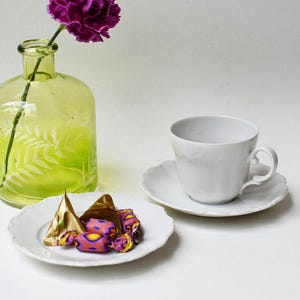 May include: A still life arrangement featuring a white teacup and saucer, a small plate with wrapped candies, and a green glass vase with a purple flower. The vase has a leaf design.