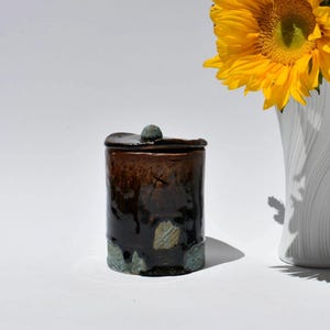 May include: A handmade ceramic jar with a lid. The jar is cylindrical with a dark brown glaze, accented with lighter brown and green hues. A small, round knob sits atop the lid. The jar is next to a white vase with a yellow sunflower.