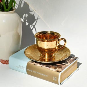 May include: A gold-colored teacup and saucer set with ornate detailing. The teacup sits on a matching saucer, both reflecting light. The set is placed on a book titled "Crime and Punishment" by Fyodor Dostoevsky.