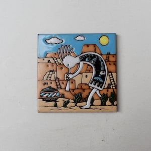 May include: Square ceramic tile featuring a Kokopelli figure in a desert landscape. The tile has a blue sky with a yellow sun and white clouds. The Kokopelli is white with black and blue details, and the background is tan and brown.