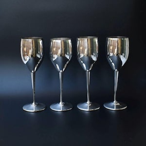May include: Four silver wine glasses with long stems and rounded bowls. The glasses are highly reflective, with the surrounding environment mirrored in their surfaces. The glasses are arranged in a row against a dark background.