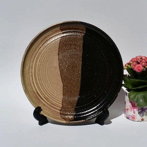 May include: A handmade ceramic plate with a unique design. The plate is divided into three sections: a light tan, a textured brown, and a glossy black. The plate is round and has a slightly raised rim. The plate is approximately 10 inches in diameter.