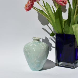 May include: A light blue ceramic vase with a textured surface and a narrow neck. Next to it is a dark blue square glass vase holding pink tulips with green leaves. The vases are on a white surface.
