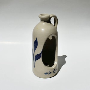 May include: A beige ceramic jug with a handle and a large, open cutout. The jug is decorated with blue leaf designs. The jug has a rounded body and a small opening at the top.