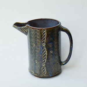 May include: A cylindrical ceramic pitcher with a dark blue and brown glaze. The exterior features a repeating pattern of horizontal wavy lines, small circular dots, and a vertical band of diagonal dashes running down the side.