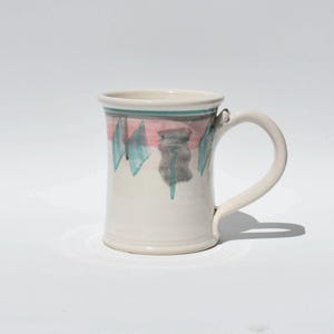 May include: White ceramic mug with a curved handle. The mug features a hand-painted design with pink, teal, and gray abstract shapes. A teal and pink band is painted around the rim. The mug is suitable for hot or cold beverages.