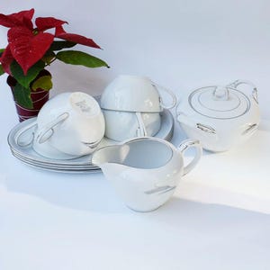 May include: A white porcelain tea set with silver trim, including a teapot, creamer, cups, and saucers. The set is arranged on a white surface, with a poinsettia plant in the background.