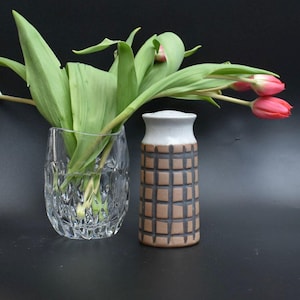 May include: A clear, cut-glass vase and a brown and gray ceramic vase with a grid pattern are displayed with fresh tulips. The tulips have green leaves and pink blooms. The vases are on a black surface.