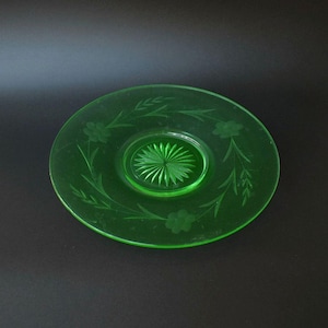 May include: A green glass plate with floral and leaf etchings. The plate has a central, oval-shaped depression with a starburst design. The rim features etched flowers and leaves. The plate is round and sits on a black surface.