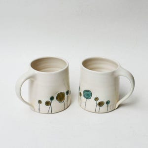 May include: Two off-white ceramic mugs with a hand-painted floral design. The mugs have a simple, rounded shape with a handle. The floral design features green and brown circular shapes with thin black stems.