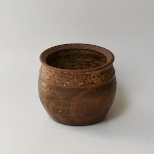 May include: A small, round, brown ceramic pot with a wide opening and a thick, rounded rim. The exterior surface features a mottled, speckled texture with darker brown swirl patterns embossed around the center of the vessel body.