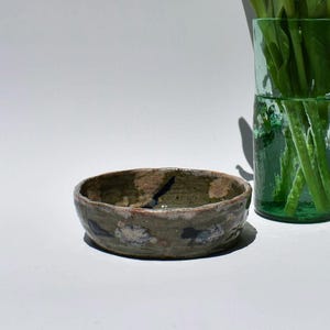 May include: A small, handmade ceramic bowl with a rustic, earthy aesthetic. The bowl features a mottled, muted color palette of greens, browns, and blues. The interior is a darker green, and the exterior has a textured, speckled appearance. The bowl is likely used for serving or display.