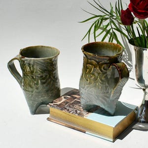 May include: Two handmade ceramic mugs with a unique, textured design in shades of green and brown. The mugs have a distinctive shape and are placed on a vintage book with gold-edged pages. A vase with red roses is in the background.