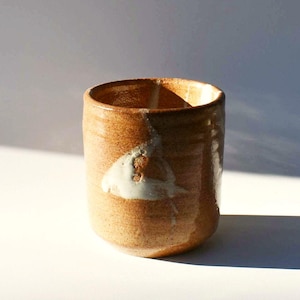 May include: A handmade ceramic cup with a cylindrical shape. The cup is primarily brown with a lighter, off-white glaze accent on the side. The interior is dark, and the cup sits on a white surface. The cup is approximately 4 inches tall.