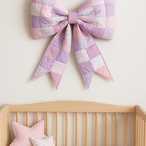 May include: A large, quilted bow in shades of lavender, pink, and white hangs above a wooden crib. The bow is made of patchwork squares. Two star-shaped pillows rest on the crib's pink sheet.