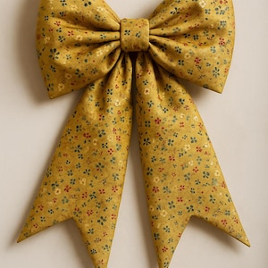 May include: A large, mustard-yellow fabric bow with a floral pattern of small red, green, and white flowers. The bow has a classic shape with two long, pointed tails. The fabric appears to be a textured cotton or linen.