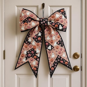 May include: A large Halloween-themed bow decoration hanging on a white door. The bow features a repeating pattern of ghosts and jack-o'-lanterns on a peach background, with black accents and trim. The bow is a festive decoration for the holiday.