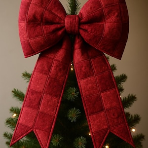 May include: A large, quilted red bow adorns a Christmas tree. The bow has a grid pattern and long, pointed tails. The tree is green with small, twinkling lights, creating a festive holiday scene.