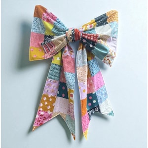 May include: A large, colorful bow made from patchwork fabric. The bow features a variety of patterns and colors, including pink, yellow, blue, and orange. The bow is tied in the center with a knot and has long, pointed tails. The bow is likely a decorative item.