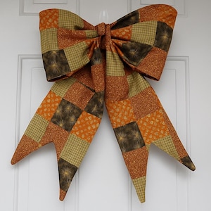 May include: A large, decorative bow made of fabric with a patchwork design. The bow features squares of orange, brown, and green fabric. The bow is hanging on a white door. The bow is approximately 30 inches wide.