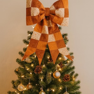 May include: A large, decorative bow in shades of orange and cream, with a checkered pattern, sits atop a Christmas tree. The tree is adorned with gold ornaments, pinecones, and warm white lights, creating a festive holiday display.