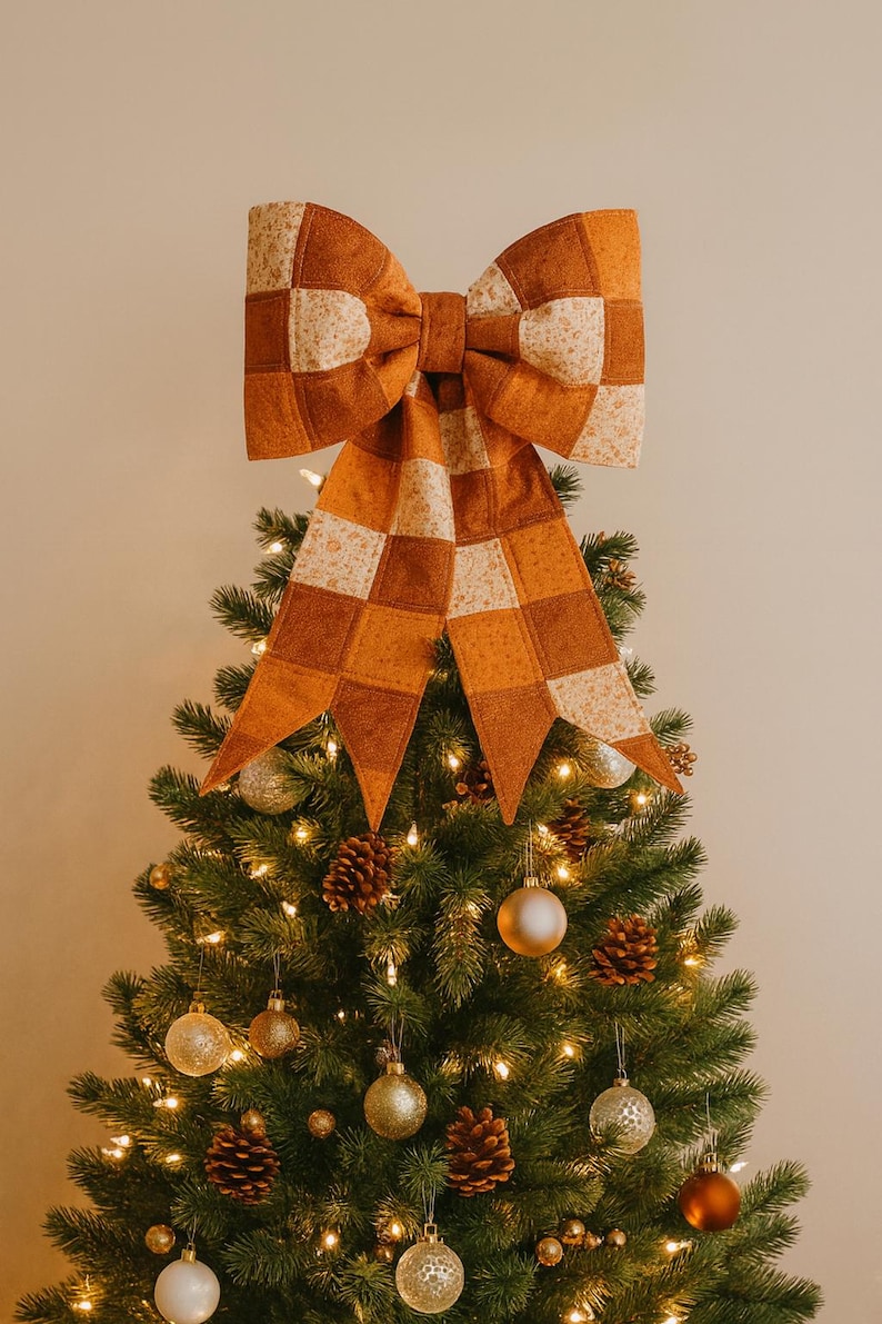 May include: A large, decorative bow in shades of orange and cream, with a checkered pattern, sits atop a Christmas tree. The tree is adorned with gold ornaments, pinecones, and warm white lights, creating a festive holiday display.
